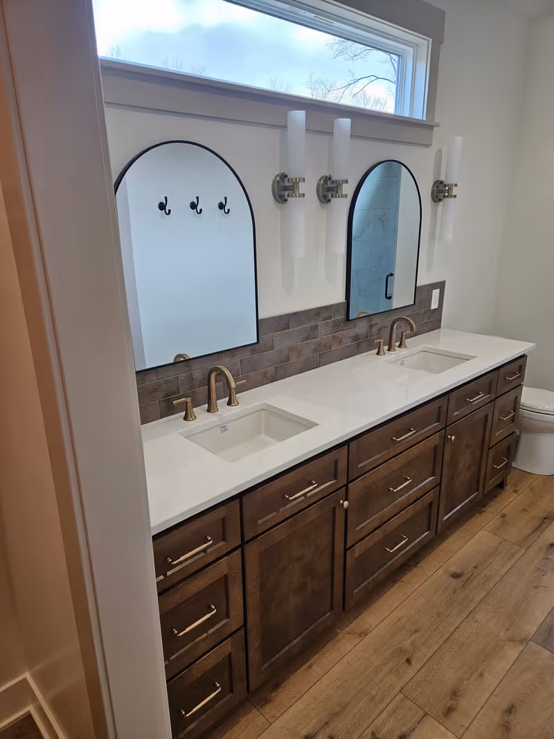 Custom double vanity with arched mirrors, dark wood cabinetry, brushed-brass faucets, and quartz countertop